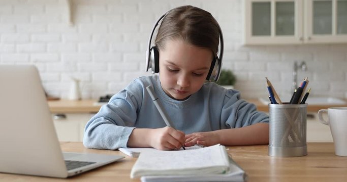 Cute kid girl wearing headphones studying at home writing notes listening audio lesson doing homework. Smiling adorable elementary school age child learning class alone sitting at table with laptop.