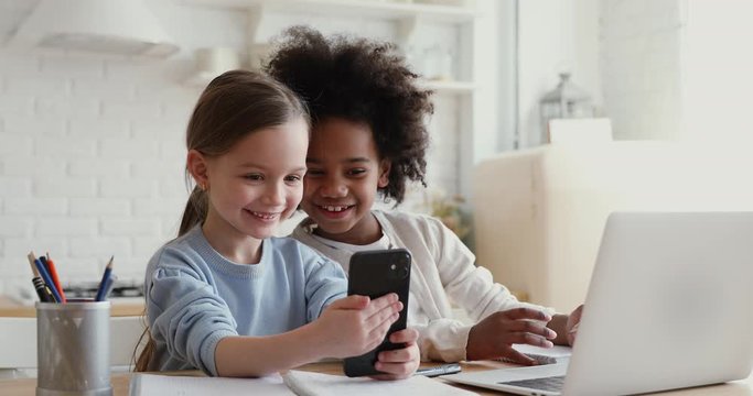 Two happy cute diverse ethnicity children using smartphone app for remote mobile education having fun playing or studying together at home. Small african and caucasian girls having fun using cell.