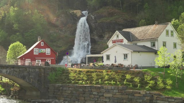 A famous Steinsdalsfossen waterfall in the village of Steine, Norway.  The wall of whitewater falling off the mountain, water spray in hanging in the air. A small cafe is nearby, welcoming tourists.