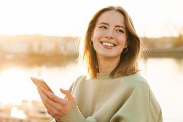 Portrait of cheerful woman using cellphone while walking on promenade