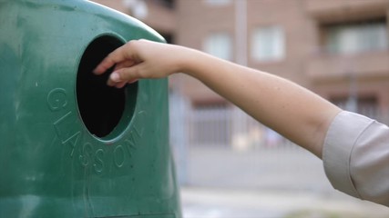 Close up of a person's hand as they dispose of broken glass into a recycling bin with the backdrop of an urban environment.