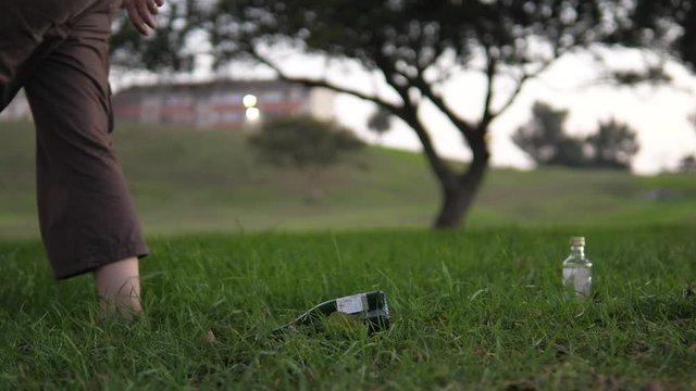 Slow-motion shot of a woman walking past, littering in a public park by dumping an empty wine bottle onto the grass.