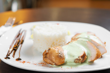 Chicken fillet with sauce is served with rice and vegetables on a white plate and a dark table. Close up.