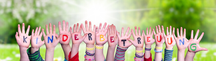 Kids Hands Holding Colorful German Word Kinderbetreuung Means Child Day Care. Sunny Green Grass Meadow As Background - obrazy, fototapety, plakaty