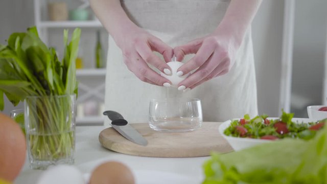 Unknown Caucasian woman breaking egg into transparent bowl. Unrecognizable young lady in apron cooking indoors. Concept of culinary, cuisine, homemade foods, healthy eating, lifestyle.