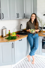 A young woman is holding a bowl of super healthy salad in her hands and smiling, she is in casual clothes in a cozy kitchen. Healthy eating concept. Vertical photo