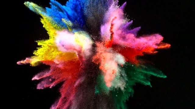 In real slow motion, a tight clump of red, blue, purple, green, yellow and black powder explodes brilliantly on a black background like a firework. Top Shot.