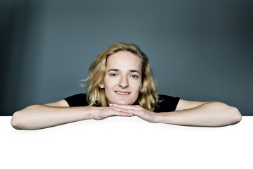 A young attractive man peeks out from above a white wall on a gray background. He laid his head in his hands. The elbows are apart.