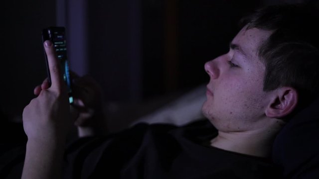 A teenage guy lying down on a bed and scrolling though the phone