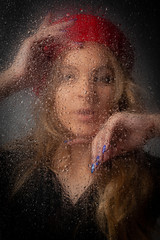 Blonde girl in a red beret behind wet glass on a gray background.