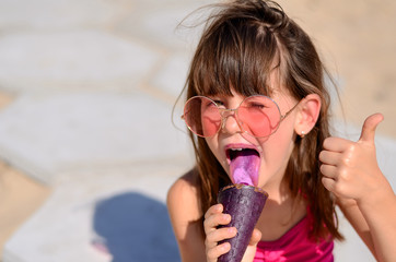 Closeup of pretty little girl eating ice cream outdoors on sunny day. Cute girl in pink swimsuit licking purple ice-cream in waffle cone. Summer, happy childhood concept. Copy space for your text