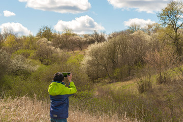 Young boy takes pictures on mobile phones of a beautiful landscape with blooming trees in early spring.