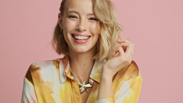 Close up view of Happy pretty blonde woman in colorful elegant costume playing with her hair and winking with eye at camera over pink background