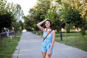 Beautiful fashionable woman is posing to the camera. Girl is wearing blue romper and white headphones. Outdoors in the park. 