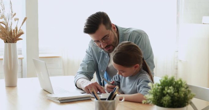 Dad helping cute small kid daughter doing homework at home. Young adult parent father or tutor explaining education teaching school girl pupil studying with daddy. Children remote education concept