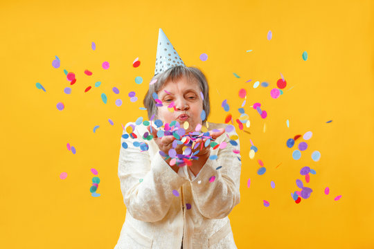 Portrait of a senior woman in studio on a yellow background. Cheerful lady blowing on confetti. Party concept