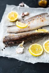 Fresh raw hake fillet on a black background. Fresh fish with lemon, garlic, salt in a spoon and peppers mix