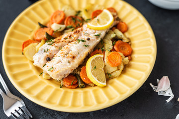 Baked hake fish in the oven with vegetables on a yellow plate on a black background. Delicious hot snacks for foodies. Selective focus