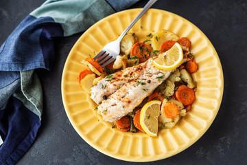 Baked hake fish in the oven with vegetables on a yellow plate on a black background. Delicious hot snacks for foodies. Selective focus