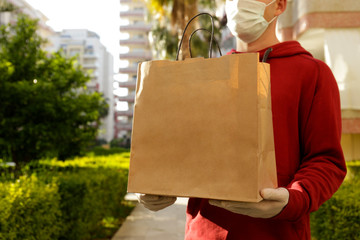 Delivery guy wearing red hoodie, disposable gloves and mask for coronavirus protection, holding blank brown paper grocery bag outside of high rise apartment building. Close up, copy space.