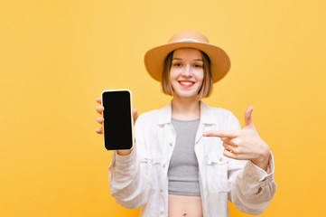 Happy lady holds a black screen smartphone in her hand and points at it with her finger, looks into the camera and smiles, wears a hat and light clothes. Copy space