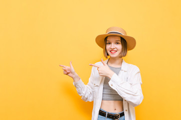 Happy lady in a light dress and hat stands on a yellow background, looks into the camera with a smile on her face and points her hands away at copyspace. Smiling girl points fingers to the empty space