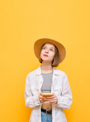 Thoughtful calm girl in hat and shirt on top stands on yellow background with smartphone in hand, looks up at copy space and thinks. Pensive lady with smartphone looking at empty space.