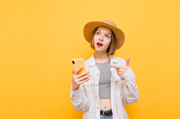 Expressive girl in hat shows finger on smartphone and looks away at copy space with surprised face on yellow background. Tourist girl with smartphone in hands and with shocked face isolated on orange