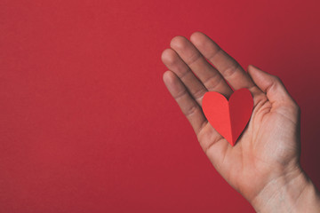 Female hand holding a red paper cut out heart on a plain red background.