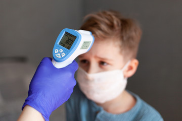 Measures the body temperature of a boy in a medical mask with an electronic thermometer.