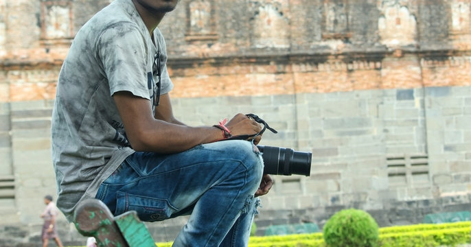 A young indian guy sitting on a bench of a park outdoor holding a DSLR camera with lens wearing t-shirt and blue jeans without face no face