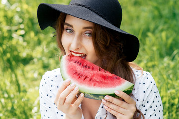 Pretty woman is eating slice of fresh watermelon wearing black hat on green grass background.