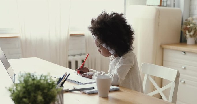Serious cute african american preschool or school child girl wearing headphones drawing studying alone sitting at home table. Mixed race kid enjoying creative activity on pandemic quarantine concept.