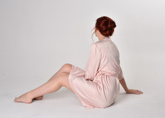 Obraz premium Portrait of a pretty girl with red hair wearing pink silk robe. sitting pose on a studio background.