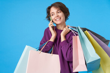 Portrait of cheerful young lovely brown haired curly lady keeping shopping bags in raised hand and smiling positively while making call, isolated over blue background