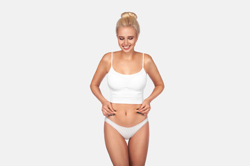 Fototapeta premium Young, healthy and beautiful blonde woman in white lingerie. Sport, fitness, diet, weight loss and healthcare concept. Slim female belly, girl holding her skin for cellulite check.