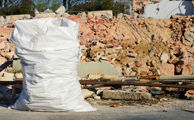 A large white plastic sack full of rubble stands in front of a demolished house with red bricks in...