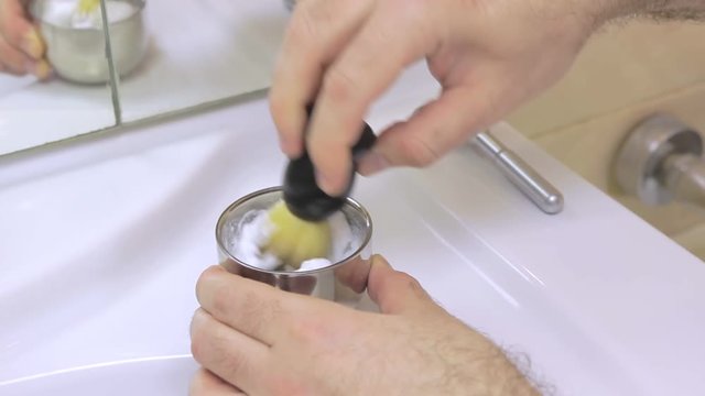 A male hand prepares shaving foam in the bathroom on a white ceramic sink. Use a classic brush and a metal chromed foam cup.