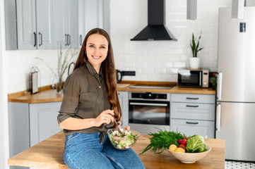 A woman in a khaki casual shirt and jeans eats a fresh wholesome salad in the kitchen from a bowl. Healthy eating concept