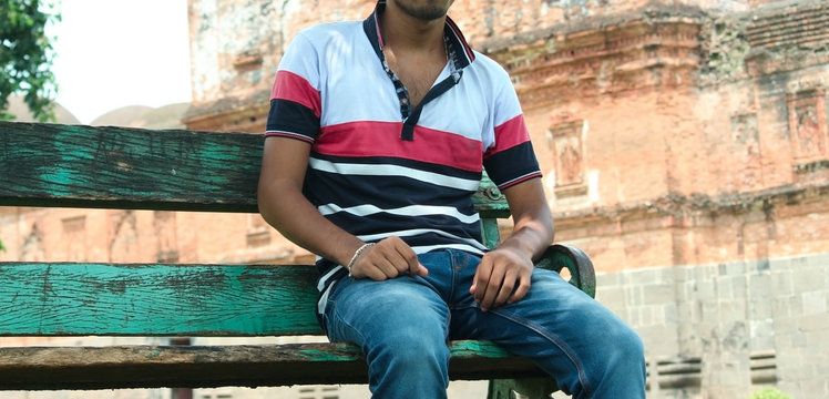 A young guy sitting on a bench in outdoor park wearing t-shirt and blue jeans and posing without face no face