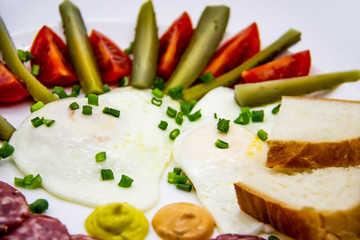 Delicious breakfast with eggs, salami, cucumbers, cherry tomatoes, slices of loaf, sauces and green onion on top on a white plate. Healthy and tasty meal for breakfast or lunch.
