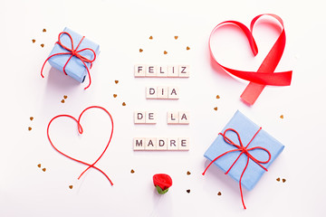 Happy Mother's Day greeting card with red hearts, blue gift boxes and golden confetti on pastel table top, overhead shot. Feliz dia de la madre text that mean Happy Mothers day