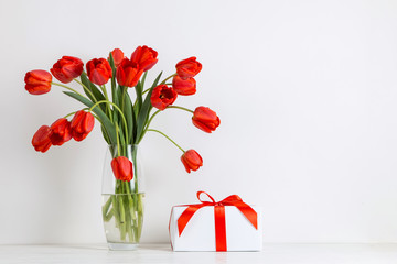 Red tulips in a vase and gift on the table, on a white background. Postcard blank.