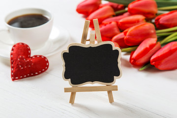 A black sign against the background of red tulips, hearts and cups of coffee on the white table. Preparing a mother's day card. Copy space.