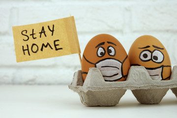 A close-up. Two eggs with painted faces in medical masks hold a poster that reads STAY HOM