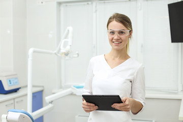 Smiling woman in white uniform holds tablet in her hands standing inside dentist office. Selective focus