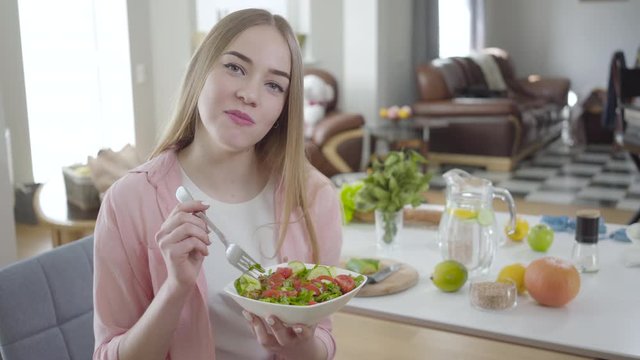 Positive young woman chewing tasty organic salad and smiling at camera. Portrait of joyful blond lady eating healthful vegetable dish. Healthy lifestyle, weight control, beauty.