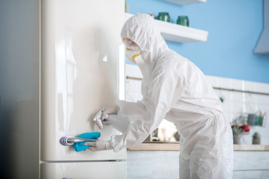 Woman in white workwear and respirator disinfecting the fridge