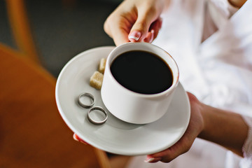 bride holds a cup with coffee and wedding rings on a saucer