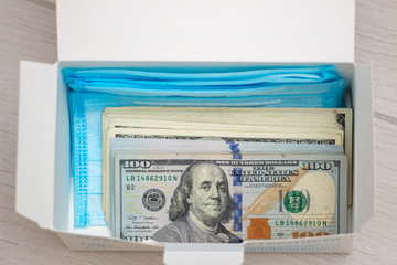 A pack of hundred-dollar bills in a box with blue disposable medical masks, close-up, top view. Medicine cost concept